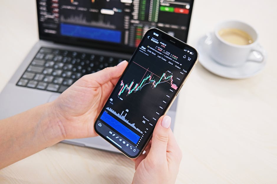 Close-up of hands holding smartphone displaying stock charts with laptop in the background on a desk.